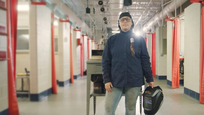 Woman wearing welding gear and holding a welding helmet